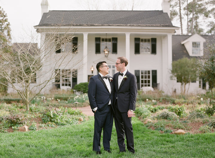 Two men in tuxedos stand facing each other and smiling in front of a large white house with black shutters, surrounded by greenery and gardens.