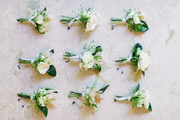 Nine small floral boutonnieres with white flowers and greenery are arranged in three rows on a light-colored, marbled surface.