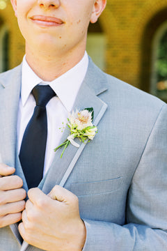 A man in a light gray suit, white shirt, and black tie adjusts his jacket. He wears a boutonniere with white flowers and greenery on his lapel. The background is softly blurred brick and arched windows.