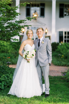 A bride in a white gown and veil stands next to a groom in a light gray suit and black tie. They are smiling outdoors in front of a white house with greenery and flowers around them.