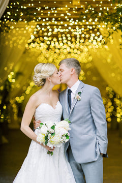A bride and groom share a kiss under a canopy of golden string lights. The bride wears a white strapless gown and holds a bouquet; the groom is in a light gray suit and tie. The background glows warmly.
