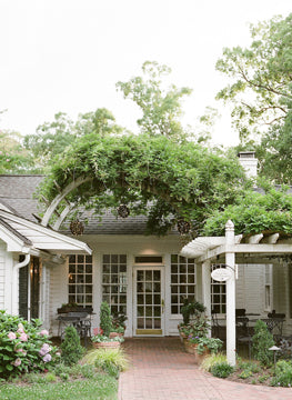 A charming white house with large windows and a vine-covered archway leads to a patio with outdoor seating, surrounded by lush greenery and blooming flowers.