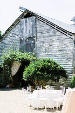 A rustic wooden barn with weathered gray siding, outdoor string lights, and white tables and chairs set up on a patio, surrounded by greenery and draped curtains at the entrance.