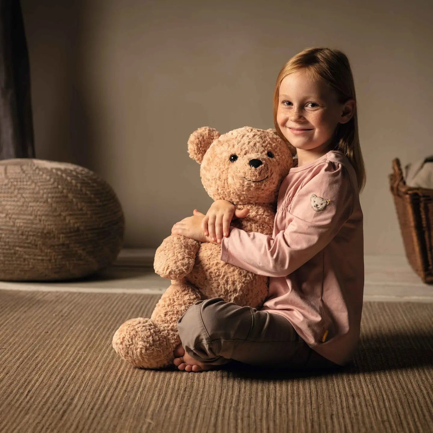 A young girl sits on a carpeted floor, smiling and hugging a large, fluffy teddy bear. She wears a light pink long-sleeve shirt and grey pants. The room has warm lighting and cozy decor.