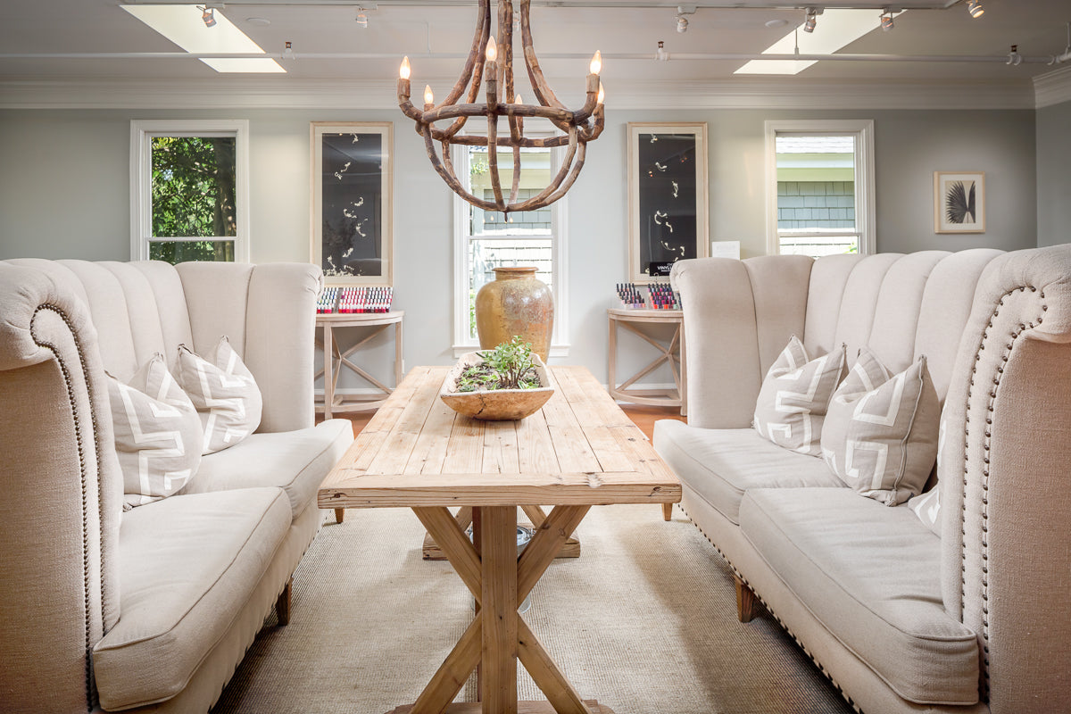 A bright, elegant room with two beige tufted sofas facing each other across a rustic wooden table. A wooden chandelier hangs above, and large windows with natural light flank decor and plants.