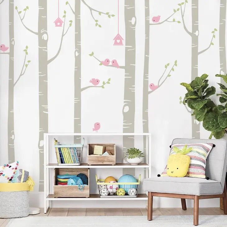 A children’s room with a wallpaper featuring illustrated trees, pink birds, and birdhouses. A white shelf holds books and toys, and a plant sits beside a chair with a pillow shaped like a pineapple.