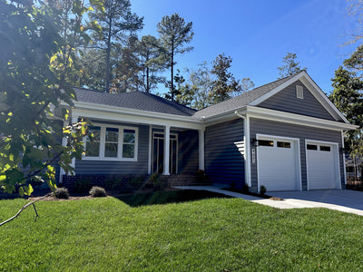 A single-story house with gray siding, white trim, and a dark shingle roof features a front porch, double garage, and a well-manicured lawn, surrounded by tall trees under a clear blue sky.