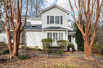A white house with gray shingles, bay windows, and green shrubs in front. Two large leafless trees and fallen leaves cover the yard, and a stone path leads to the entrance on a partly cloudy day.