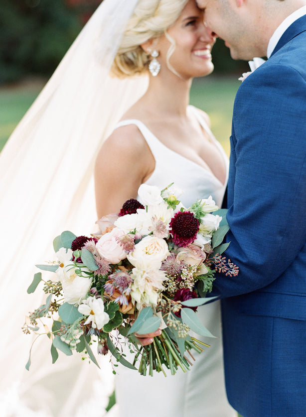 A bride in a white dress holding a bouquet of pastel and burgundy flowers smiles closely with a groom in a blue suit outdoors, both facing each other and touching foreheads.