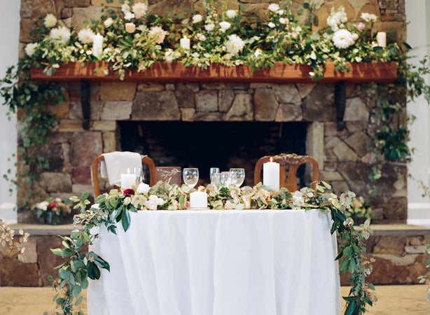 A round table with a white tablecloth is decorated with greenery, white flowers, candles, and glassware. Behind it is a stone fireplace adorned with more flowers and candles, creating a romantic setting.