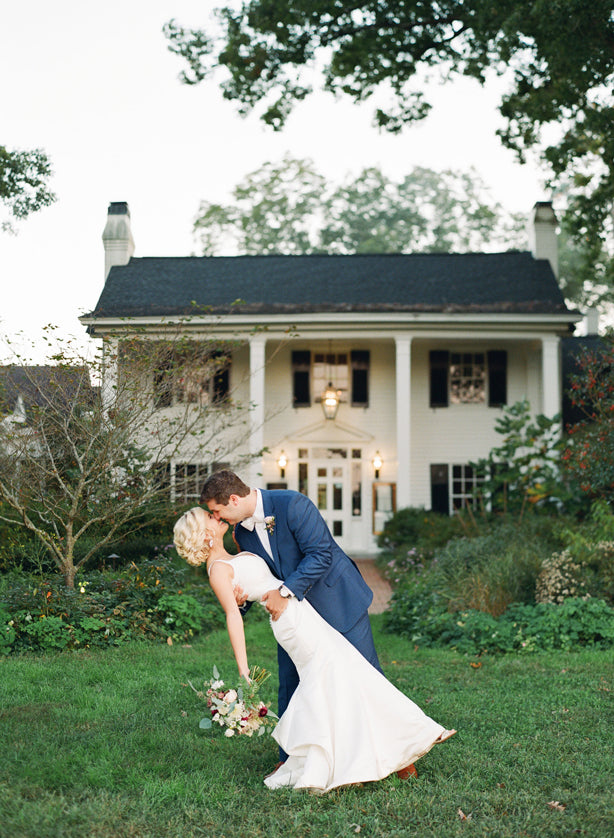 A bride and groom share a kiss as the groom dips the bride in front of a large, elegant white house with black shutters, surrounded by green lawn and lush garden landscaping.