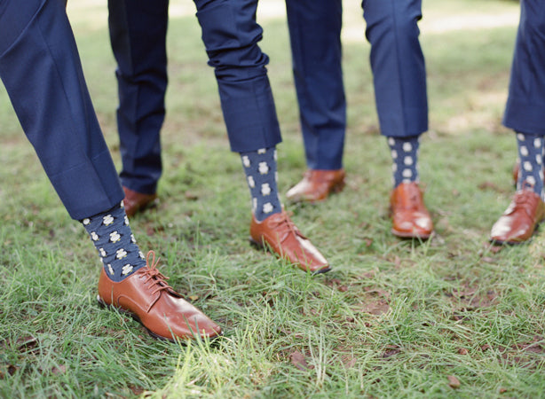 Three people wearing blue suits and brown dress shoes stand on grass, showing off matching blue socks with white floral patterns. Only their legs and feet are visible.