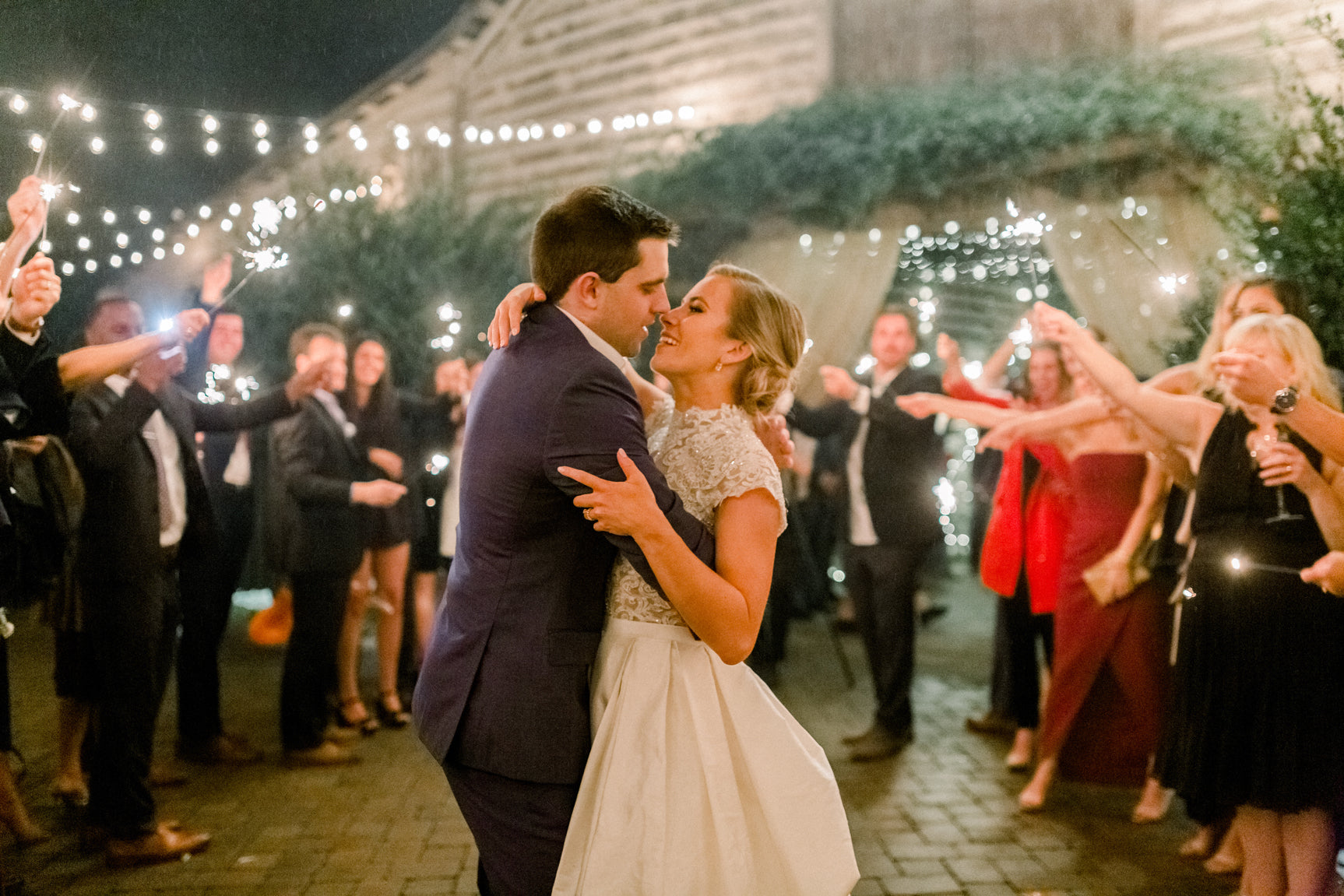 A bride and groom share a romantic first dance outdoors at night, surrounded by guests holding sparklers and string lights, creating a festive and joyful wedding celebration atmosphere.
