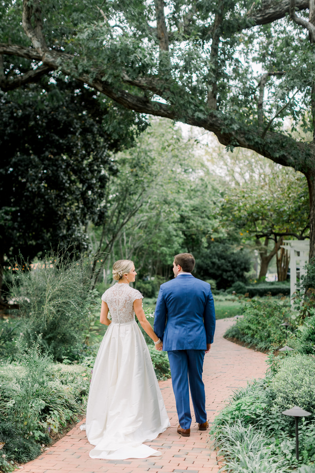 A bride in a white dress and a groom in a blue suit walk hand-in-hand down a brick path surrounded by lush greenery and trees in a garden setting.