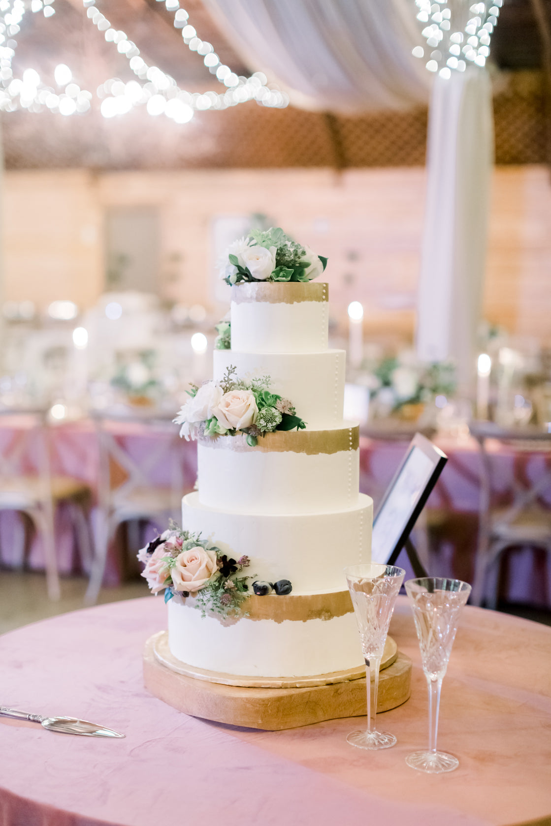 A four-tier white wedding cake with gold bands and floral decorations sits on a round table with two champagne flutes. The background features pink tablecloths, candles, and draped string lights.