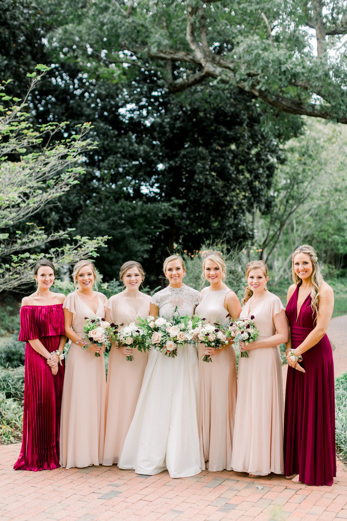 A bride in a white gown stands outdoors with six bridesmaids. The bridesmaids wear pale pink or burgundy dresses and hold bouquets. Trees and greenery form the background on a brick path.