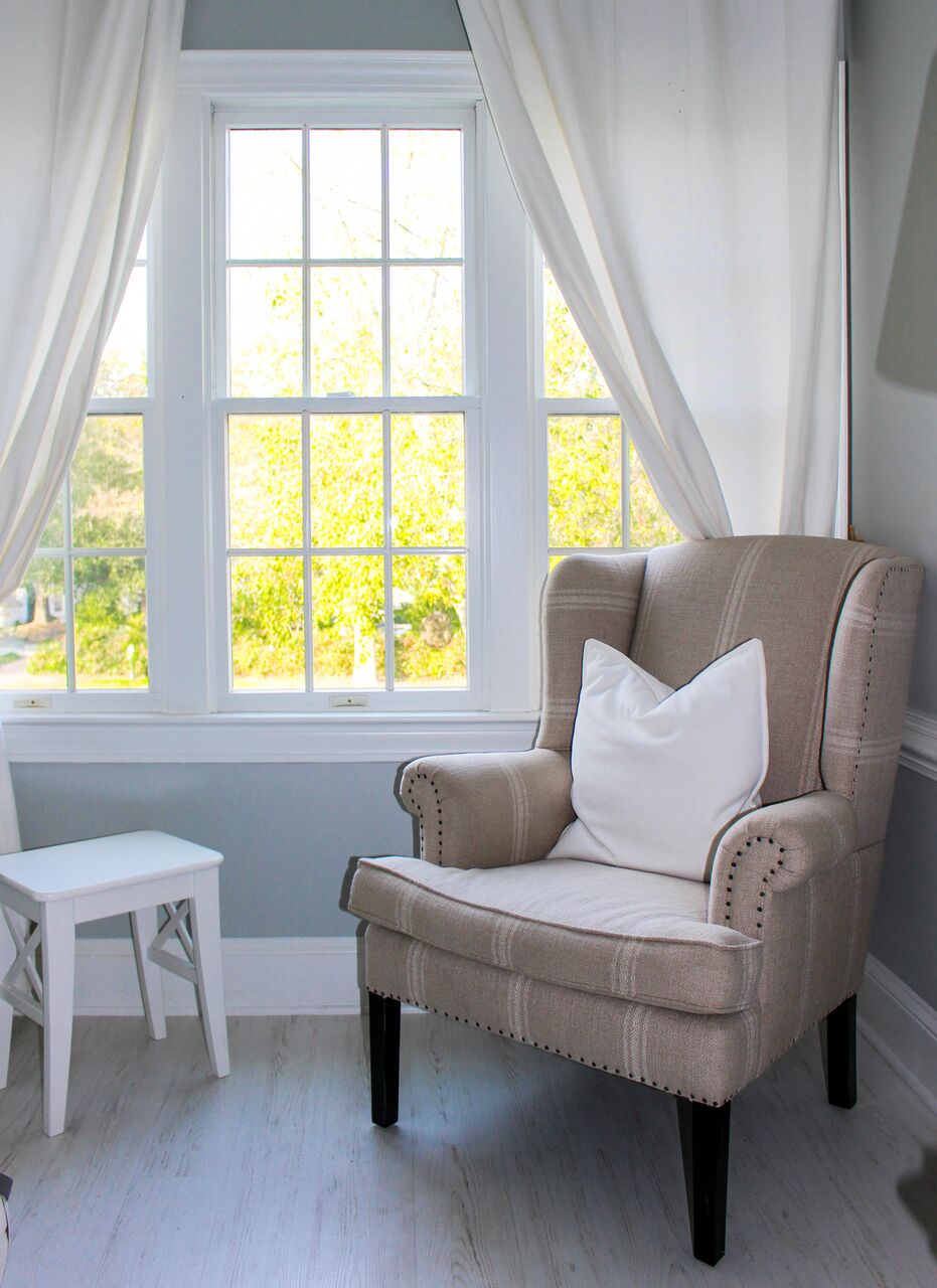 A beige armchair with a white pillow sits by a bright window with white curtains. A small white side table is nearby. Sunlight streams in, illuminating the cozy, light-colored room.