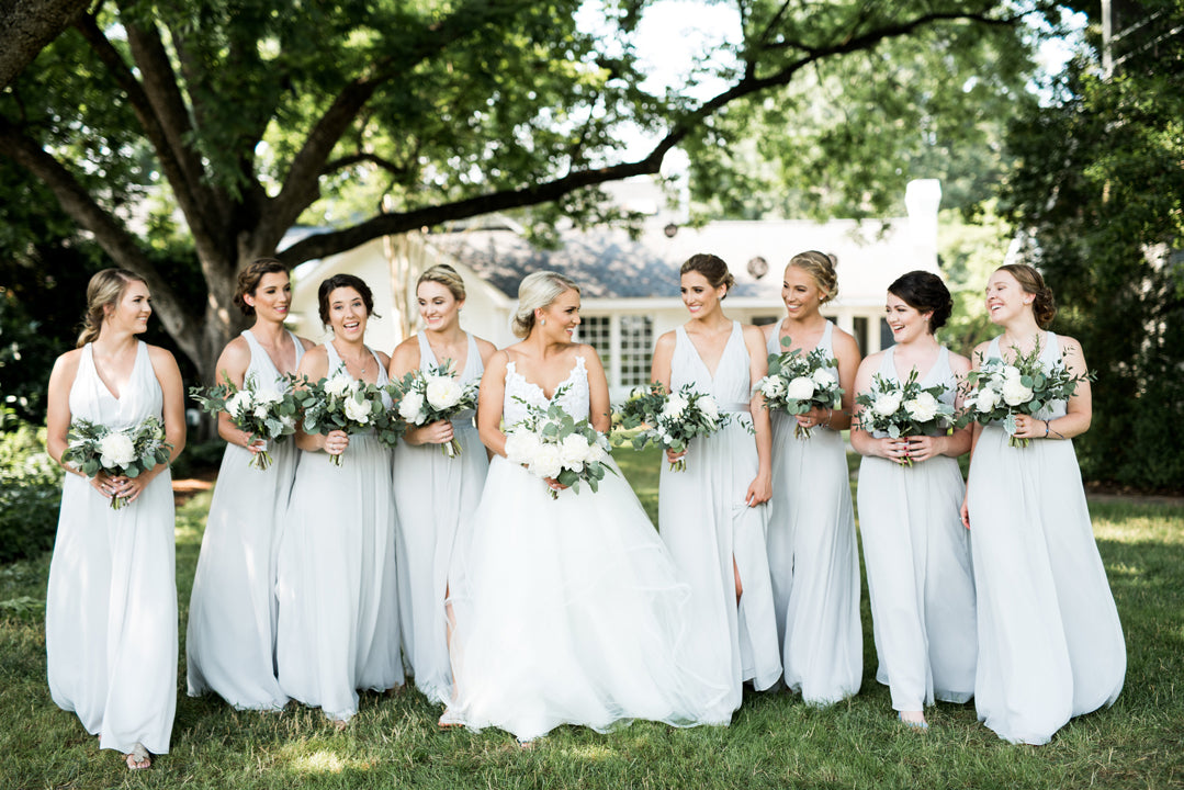 A bride in a white dress stands outdoors, smiling with her eight bridesmaids in light gray dresses, all holding bouquets, under large green trees with a house in the background.