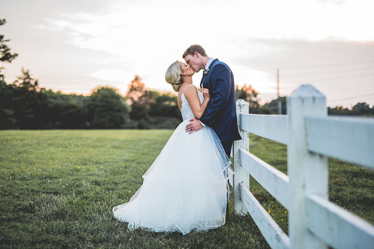 A bride and groom share a kiss beside a white wooden fence in a grassy field at sunset. The bride wears a white gown and the groom is in a dark suit. Trees and a soft, colorful sky are in the background.
