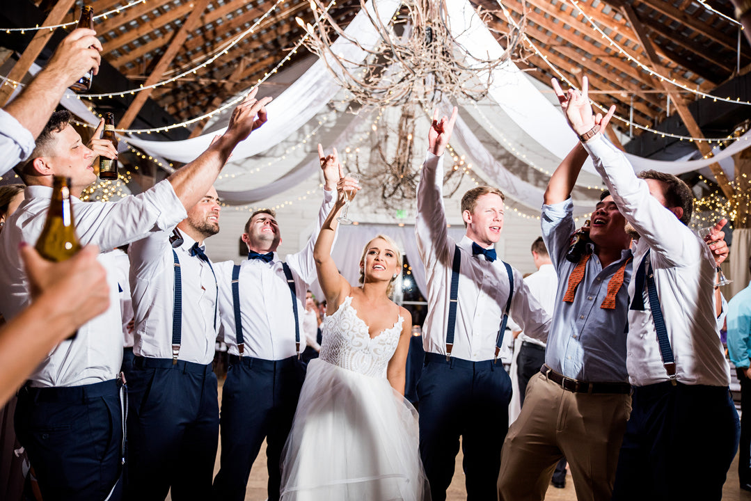 A bride in a white dress dances joyfully with a group of men in suspenders and bow ties under string lights and draped fabric at a lively wedding reception. Everyone is smiling and pointing upward.