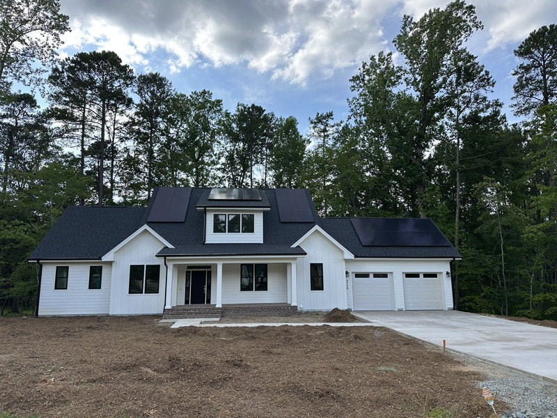 A modern white house with black roof shingles and solar panels sits in front of tall trees. The front yard is mostly bare dirt with a concrete driveway and walkway leading to the entrance.