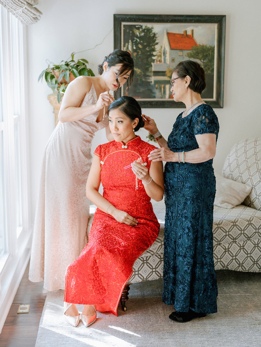 A woman in a red traditional dress sits as two women, one in a pale gown and one in a navy lace dress, help her with jewelry and hair, in a bright room with a window and a framed painting on the wall.