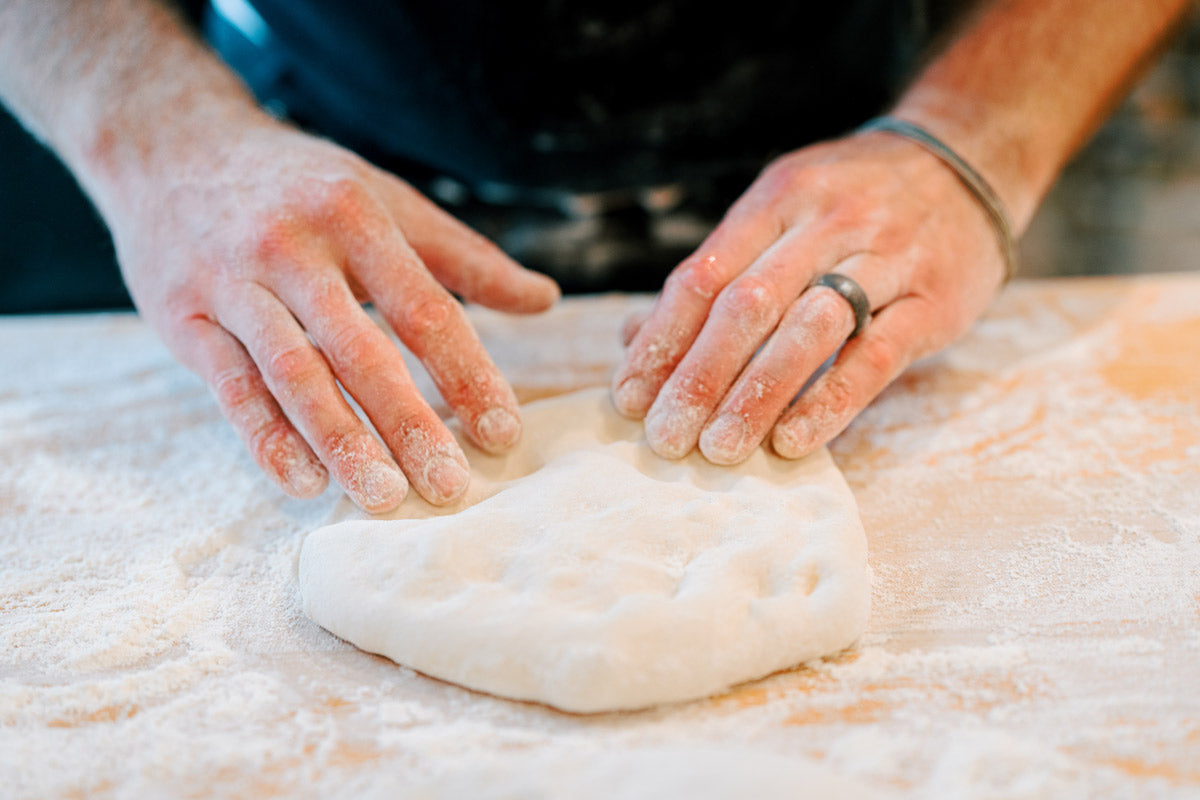 Hands pressing and shaping a mound of flour-dusted dough on a floured surface, preparing it for baking. The person is wearing a dark shirt and a ring on one finger.
