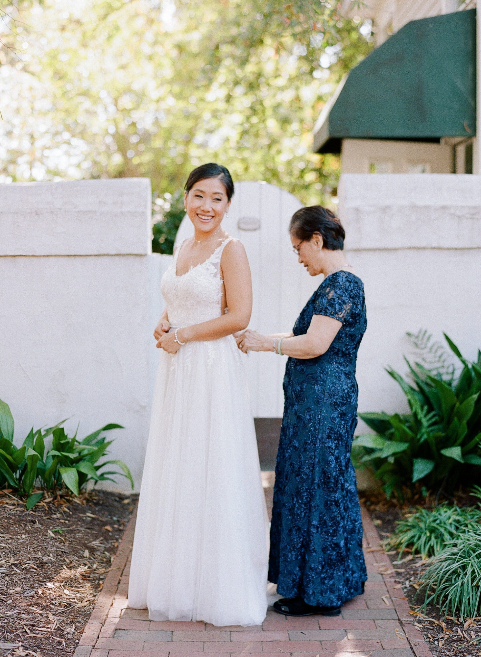 A bride in a white gown smiles at the camera while an older woman in a navy lace dress fastens the back of her dress outdoors, near a white wall and green plants.