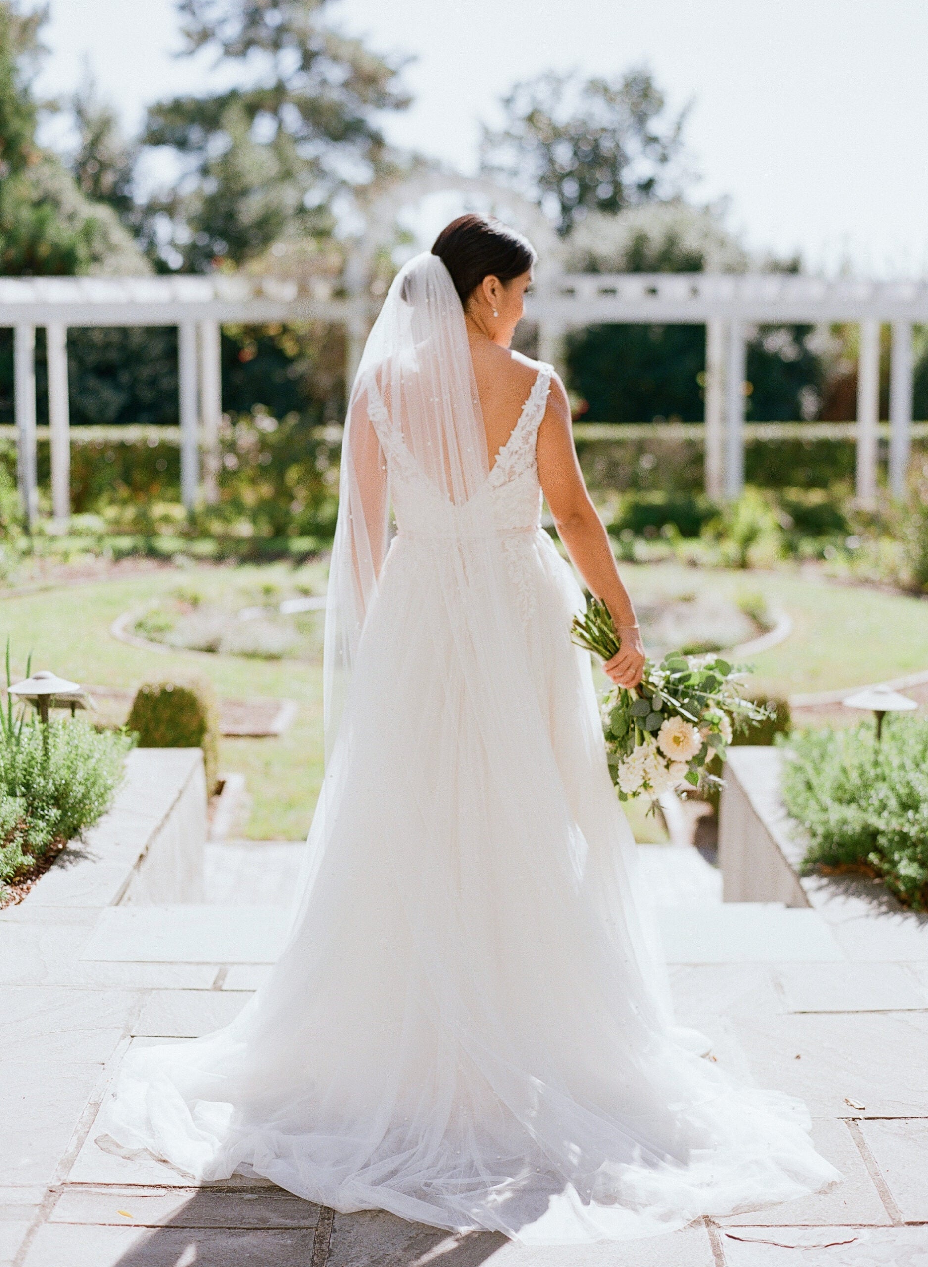 A bride in a white gown and veil stands outdoors, holding a bouquet of flowers, with her back to the camera. She is on stone steps in a sunlit garden with greenery and a white pergola in the background.