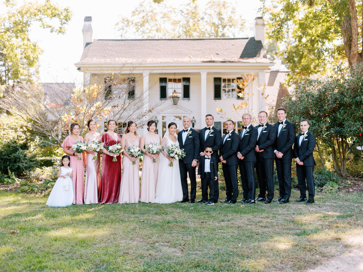 A wedding party poses outdoors in front of a white house. Bridesmaids in pink and red dresses hold bouquets, while groomsmen in black suits stand beside them. Two children, a girl in white and a boy in a suit, stand at each end.