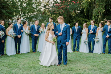 A bride and groom stand together on grass, smiling at each other, surrounded by their wedding party dressed in blue suits and light blue dresses, holding bouquets, with trees in the background.