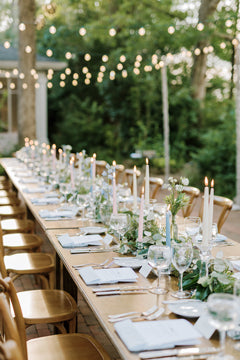 A long outdoor banquet table is elegantly set with glassware, plates, menus, and tall candles. String lights hang above, and greenery decorates the table, creating a romantic and festive atmosphere.