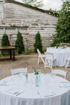 A round table set for an outdoor event with white tablecloth, glassware, and a small floral centerpiece in a blue vase; rustic wooden building and greenery in the background.