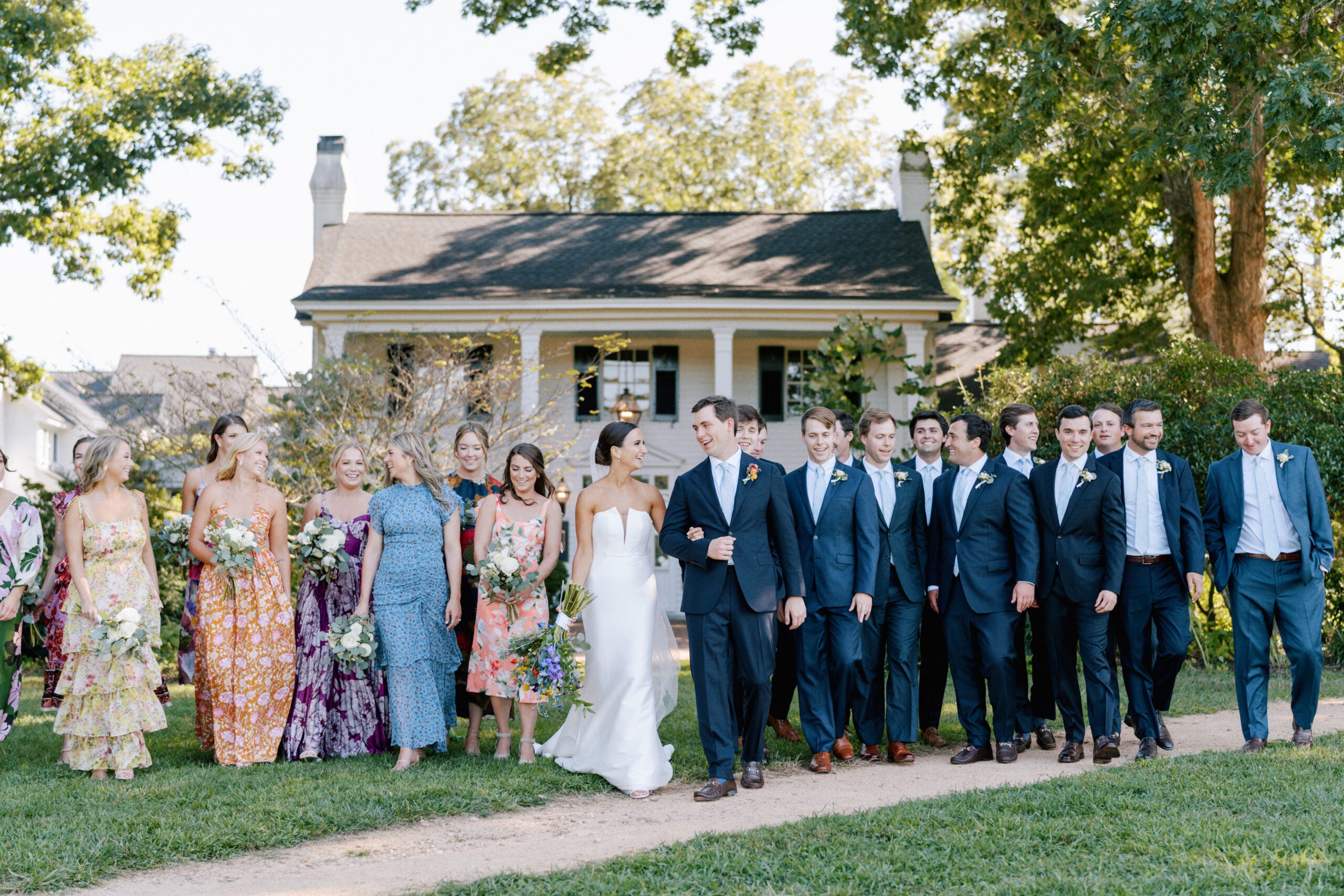 A bride and groom walk outside with their wedding party, who are dressed in colorful dresses and dark suits, in front of a house surrounded by green trees and lawn on a sunny day.