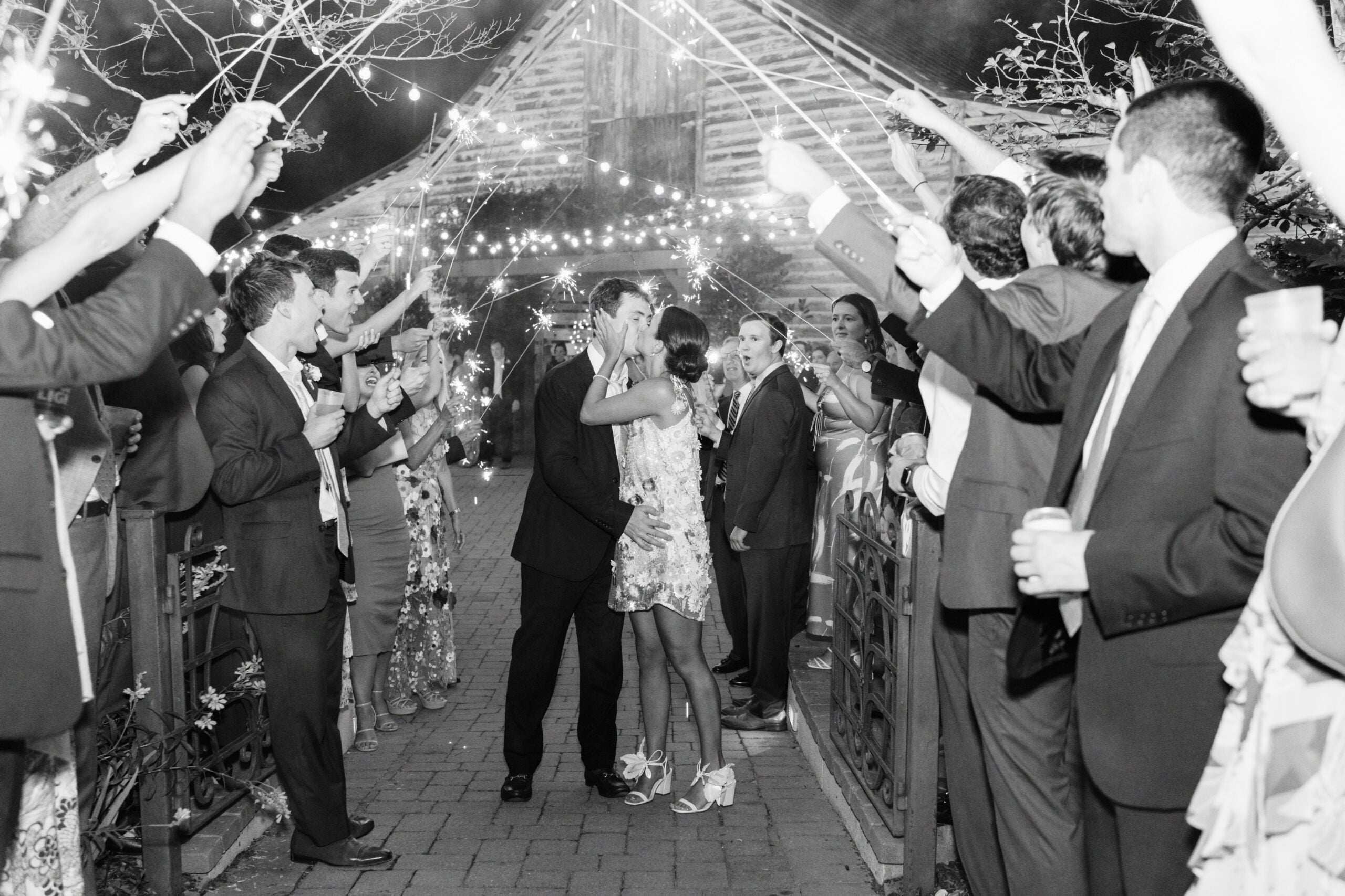 A couple kisses in the center of a pathway while guests on both sides hold up sparklers, creating a festive, celebratory atmosphere outside a rustic barn at night.