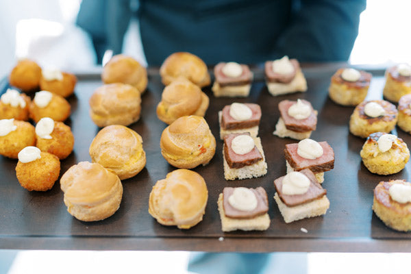 A tray of assorted bite-sized appetizers, including round pastry puffs, bread squares topped with meat and sauce, and golden fried balls. The tray is being held by a person in a dark outfit.