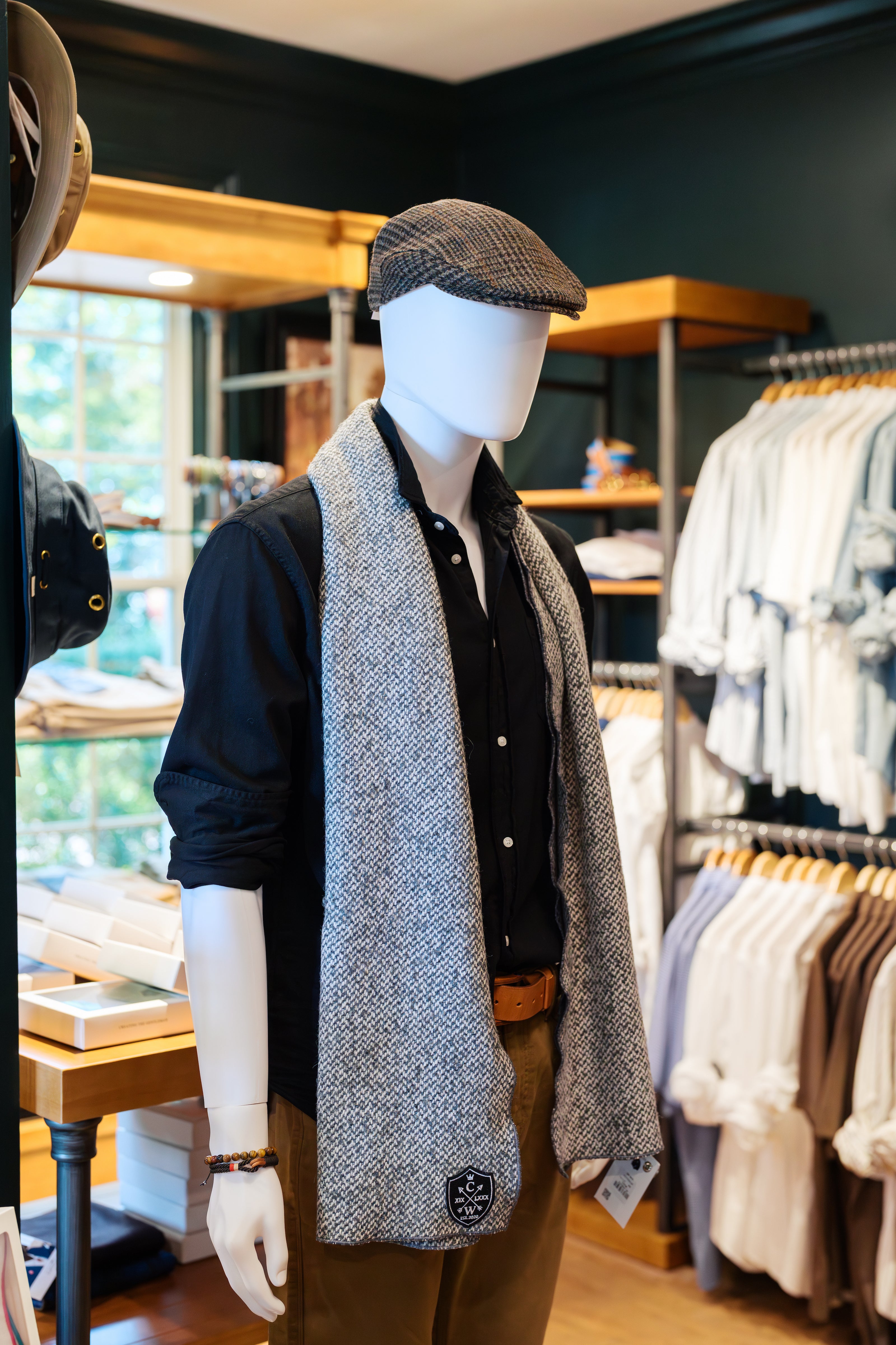 A mannequin dressed in a black shirt, tan pants, gray patterned scarf, and flat cap stands in a stylish clothing store surrounded by neatly displayed shirts and shelves.
