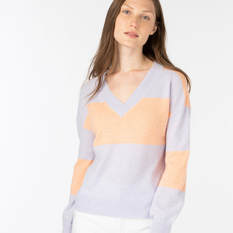 A woman with long brown hair wears a pastel purple and peach striped V-neck sweater and white pants, standing against a plain white background. She is looking slightly to the side with a neutral expression.