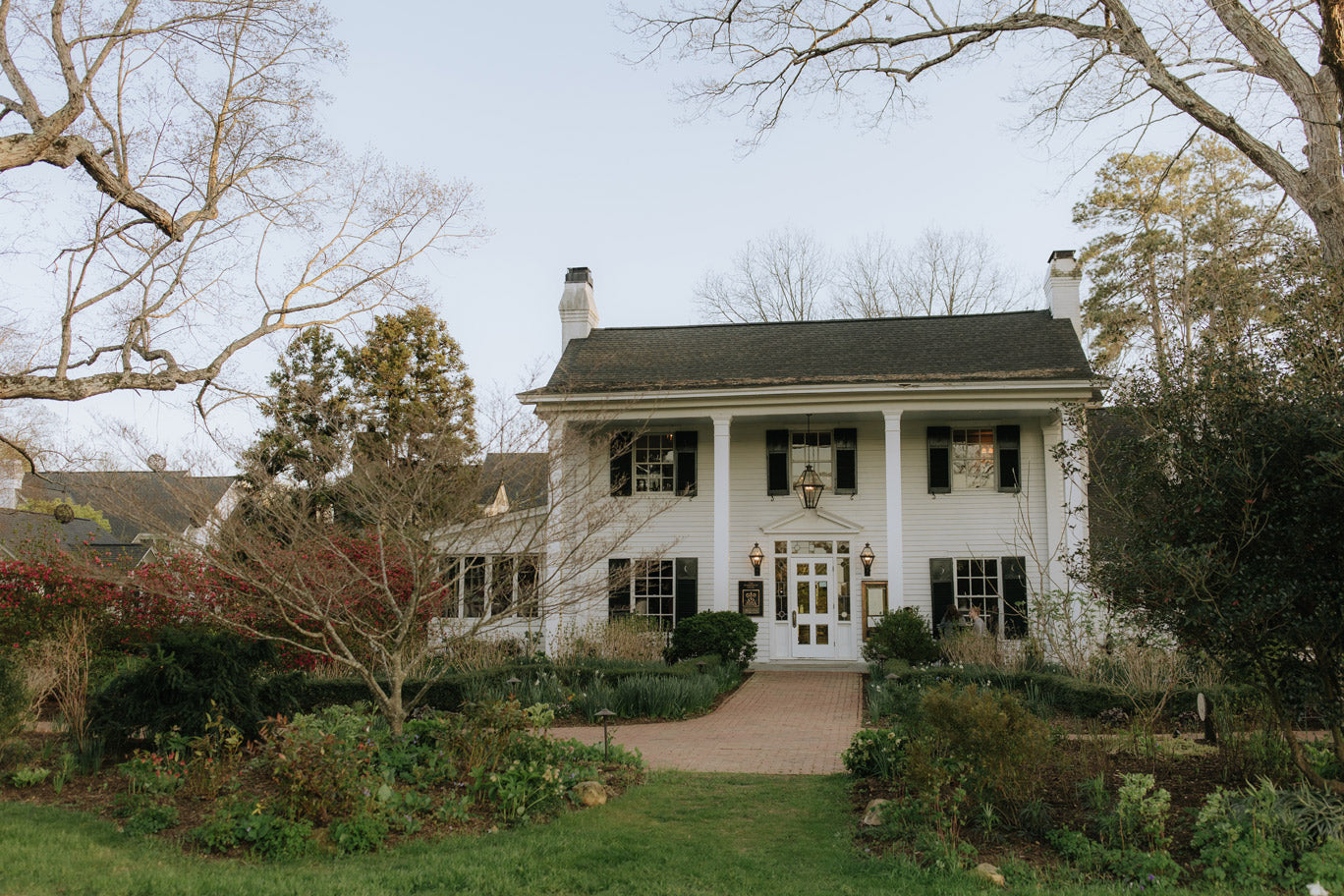 A large white two-story house with black shutters and columns sits behind a brick walkway, surrounded by gardens and bare trees under a clear sky.