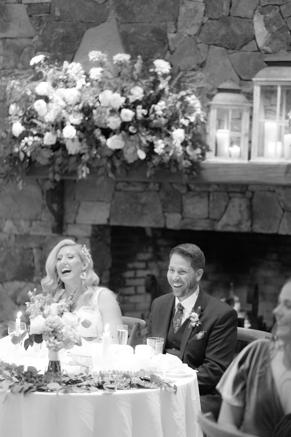 A bride and groom sit together at a decorated table, smiling and laughing during a wedding reception. Behind them is a stone fireplace with candles and a large floral arrangement.