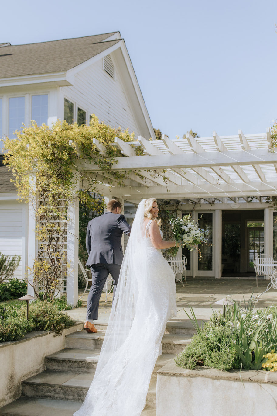 A bride in a white dress and veil holds a bouquet and looks back while walking up steps with a groom in a suit toward a white house with a pergola covered in greenery on a sunny day.