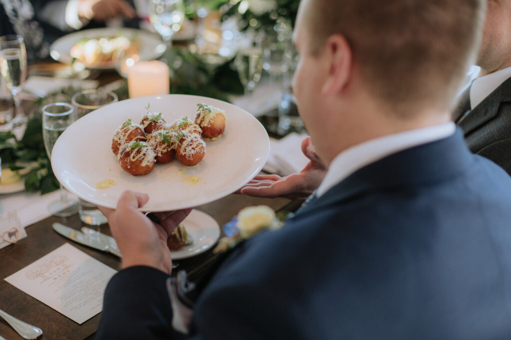 A person in a suit holds a white plate with six garnished, round appetizers at an elegant table set with glasses, greenery, and a lit candle.