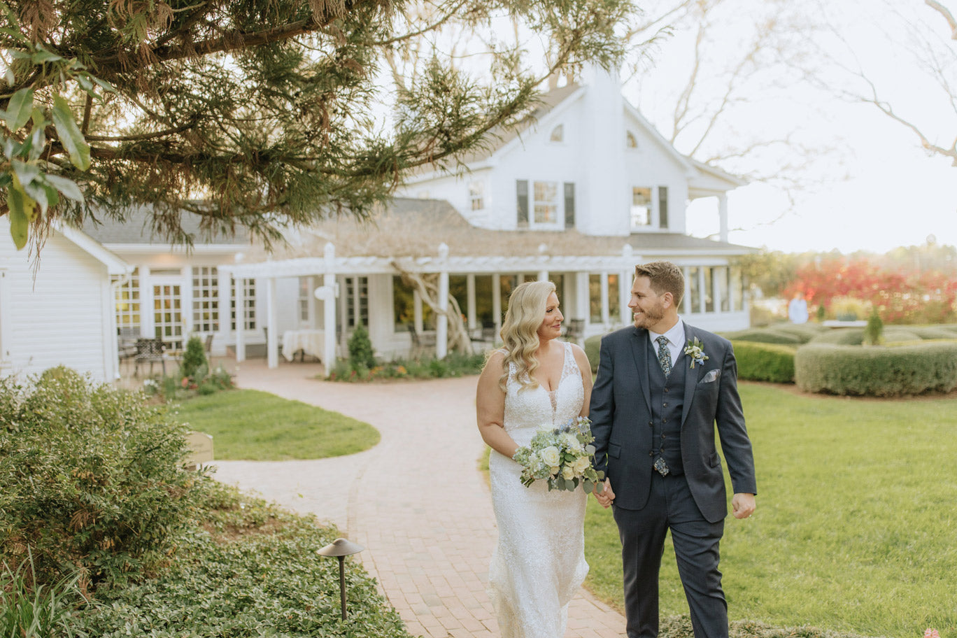 A bride in a white dress and a groom in a navy suit walk hand in hand on a brick path outside a white house with gardens, smiling at each other on their wedding day.