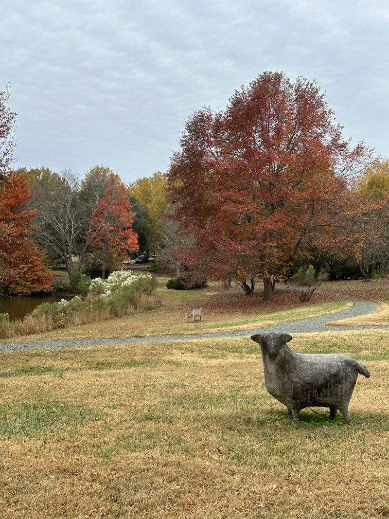 A stone dog sculpture stands on a grassy field near a winding gravel path, with colorful autumn trees and a small pond in the background under a cloudy sky.
