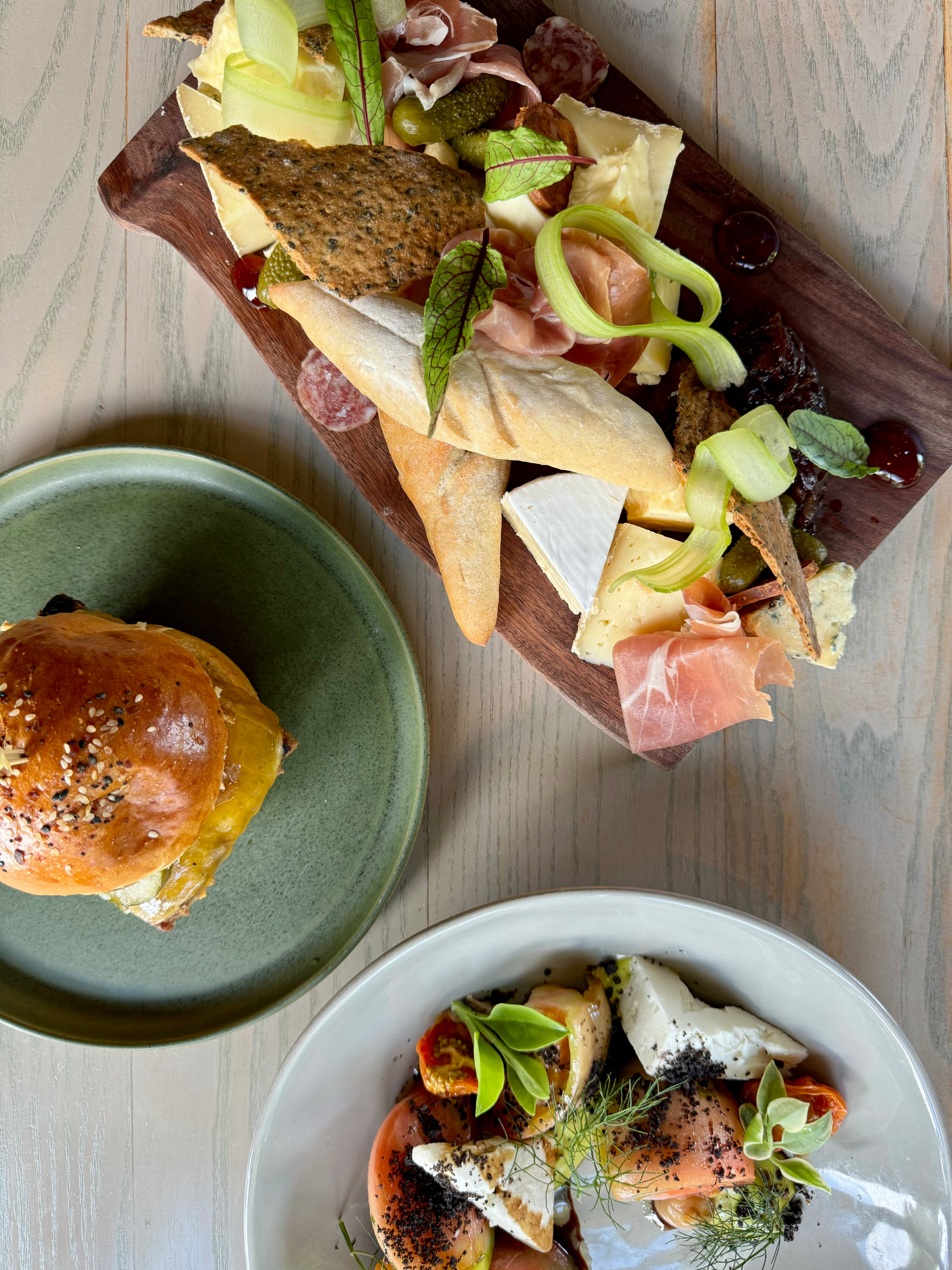 A wooden board with assorted cheeses, cured meats, crackers, and pickles next to a sandwich on a green plate and a salad with tomatoes, burrata, herbs, and edible flowers on a white plate, all on a light wooden table.