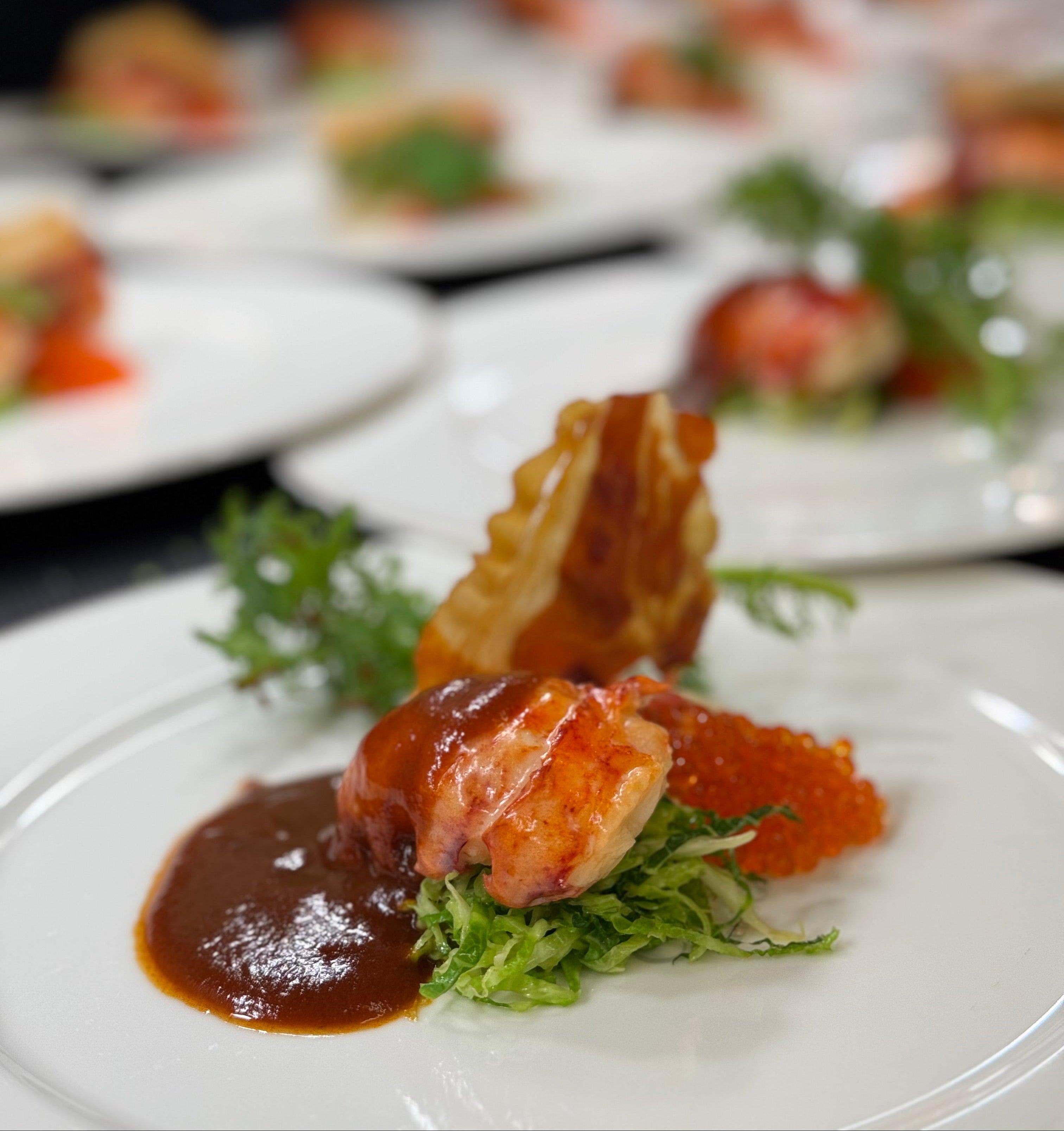 A gourmet dish featuring a piece of lobster with a rich brown sauce, orange fish roe, crispy garnish, and fresh greens, beautifully plated on a white dish with other similar plates blurred in the background.