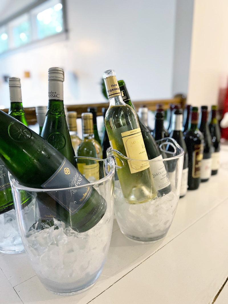 Several bottles of white wine are chilling in clear ice buckets on a table, with additional bottles of red and white wine lined up in the background. The setting appears bright and modern.