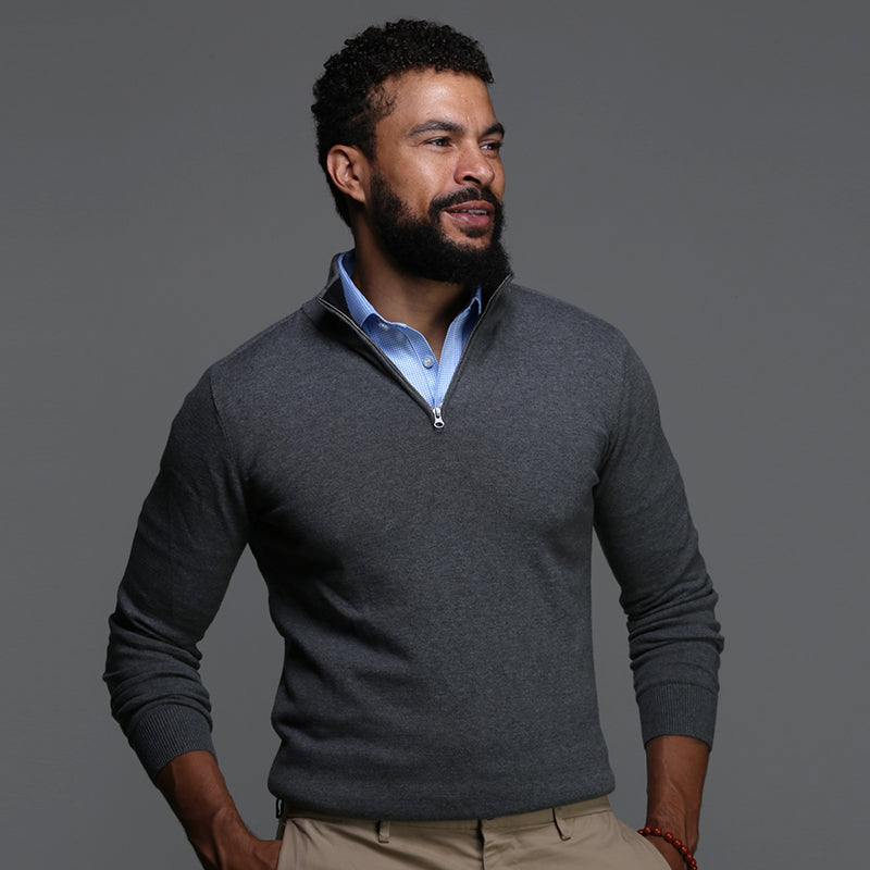 A man with short curly hair and a beard wears a gray quarter-zip sweater over a light blue button-down shirt and beige pants, standing against a plain gray background and smiling slightly while looking to the side.