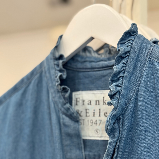 Close-up of a blue denim shirt with ruffled edges on the collar, hanging on a white hanger. The label inside reads