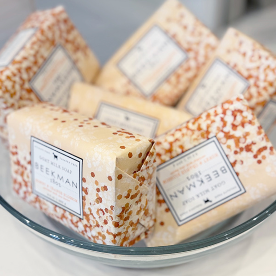 A glass bowl filled with several rectangular bars of Beekman 1802 goat milk soap, each wrapped in patterned cream and orange paper with labeled packaging.